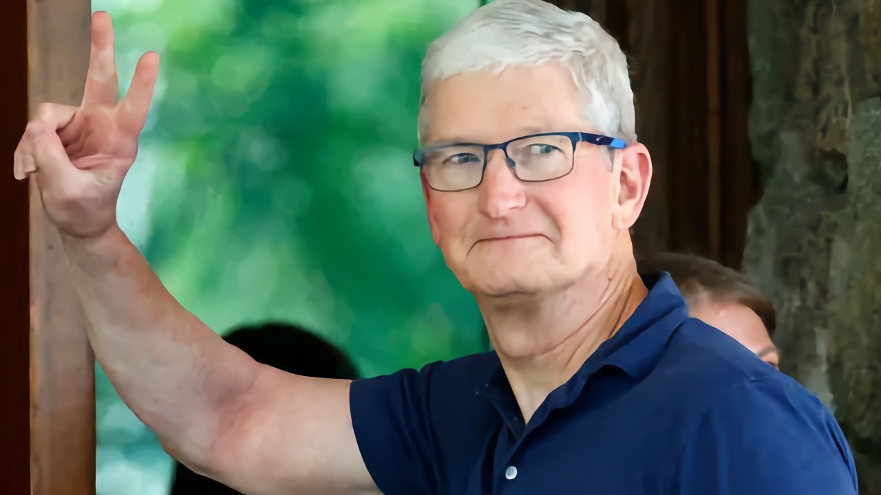 Man with glasses and white hair, wearing a blue shirt, holding up two fingers in a peace sign, standing against a blurred green background.