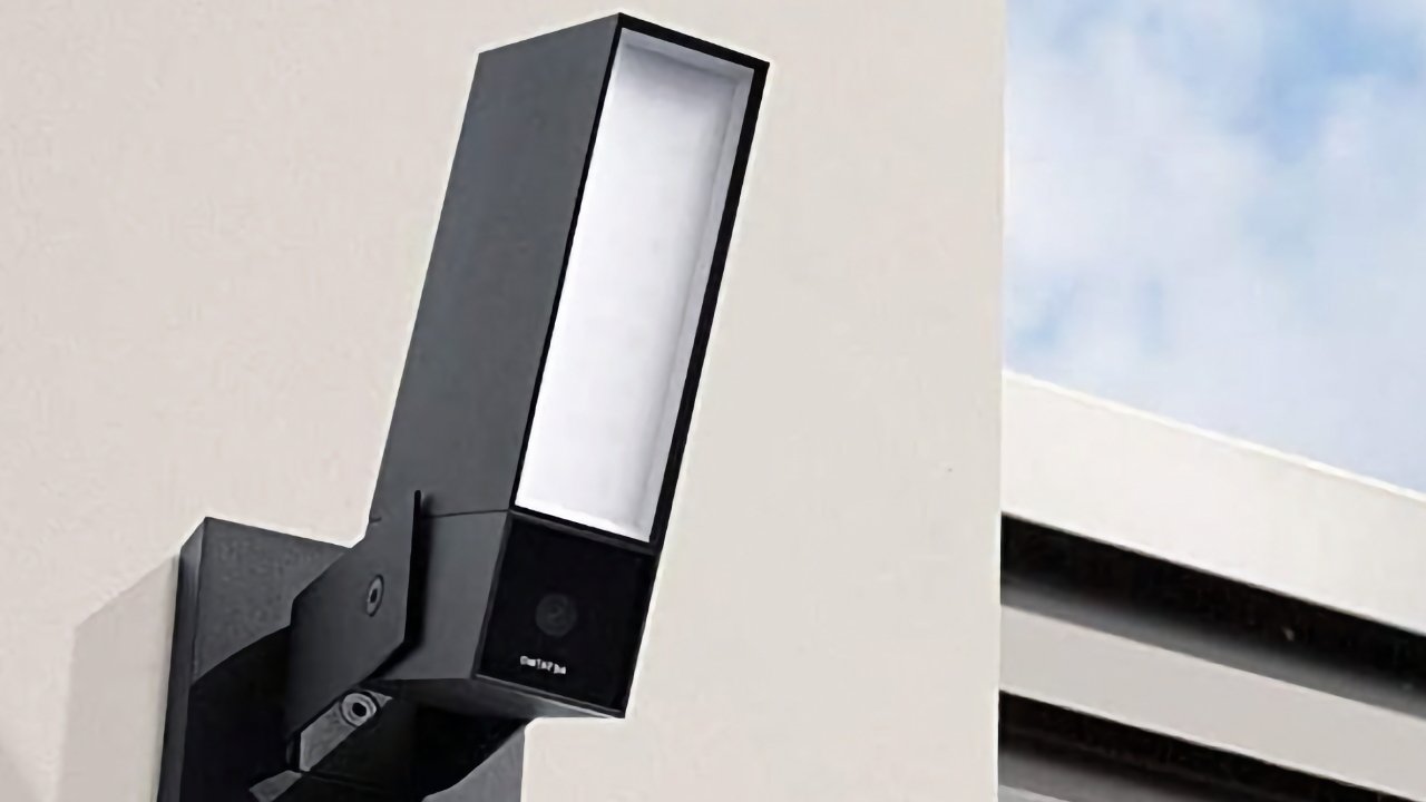 Wall-mounted modern security camera with sleek design, featuring a rectangular lens against a beige background and a partial view of the sky.