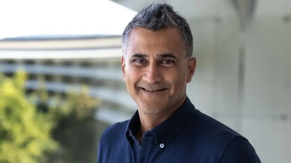 A smiling person with short gray hair wearing a dark shirt, standing outdoors with a blurred modern building and greenery in the background.