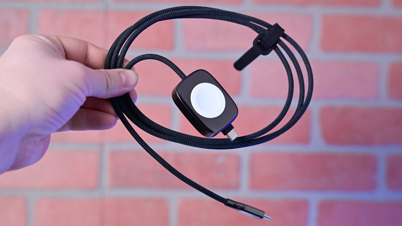 Hand holding a black charging cable with a USB-C connector and a magnetic charging pad against a blurred brick background.