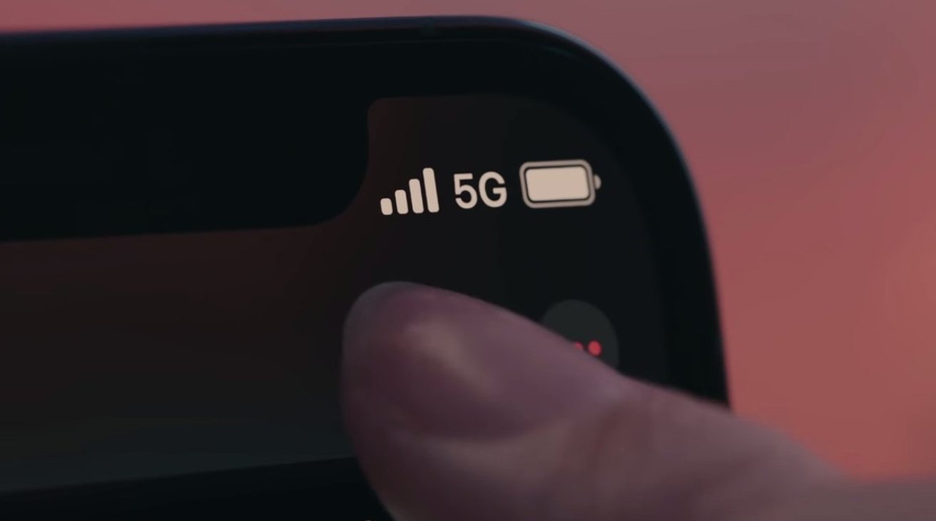 Close-up of a smartphone screen corner displaying 5G signal, battery icon, and a fingertip touching the screen on a blurred background.