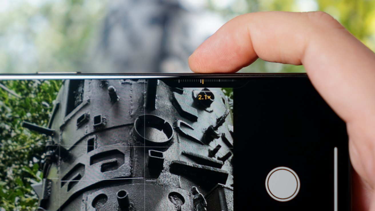 Person holds phone, zooming in on a detailed, cylindrical metal sculpture outdoors, surrounded by greenery.