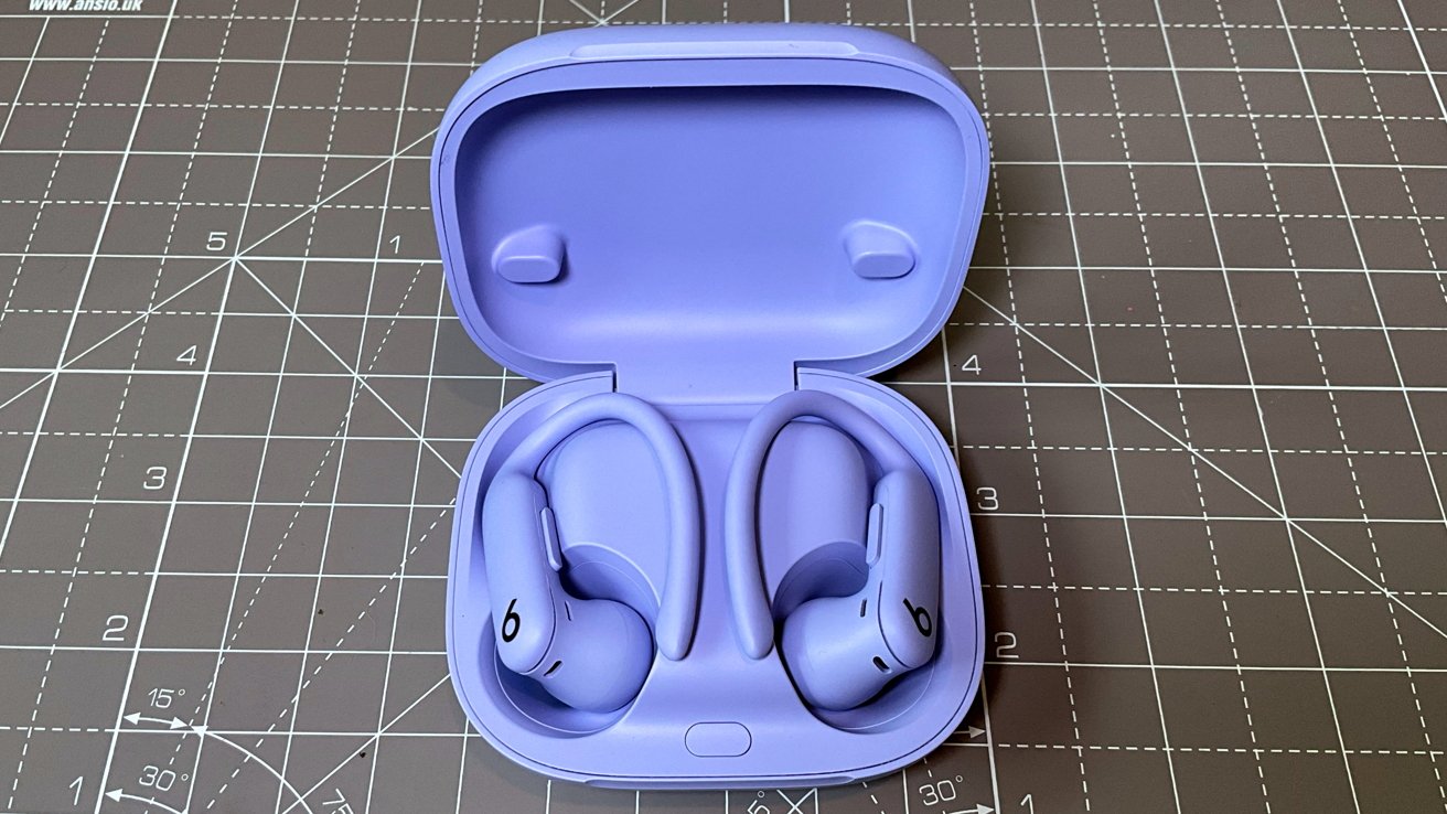 Purple wireless earbuds with ear hooks in an open matching case, resting on a gridded cutting mat.