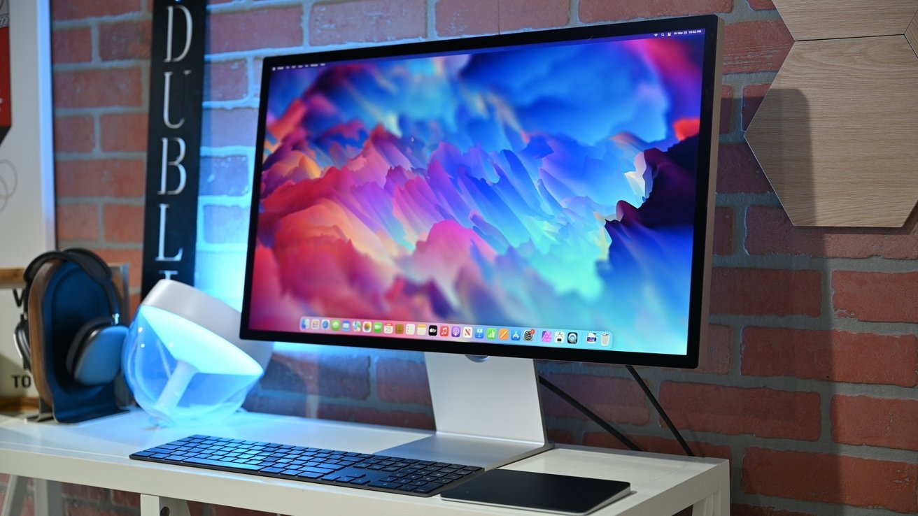 Apple Studio Display displaying colorful abstract art on a white desk with a black keyboard, headphones, and a lit spherical lamp against a brick wall.