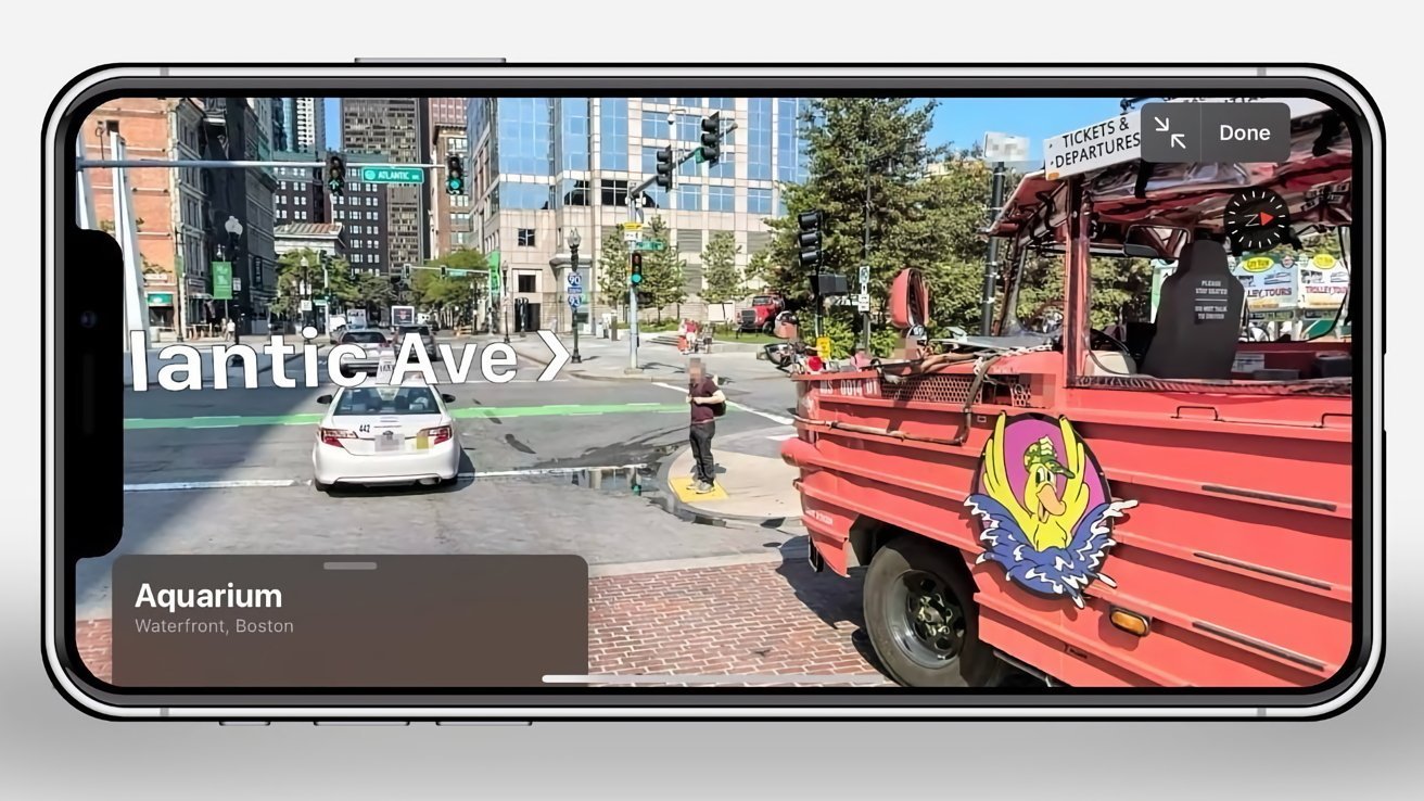 Smartphone displaying street view with a red tour vehicle, white car, city buildings, trees, and signs reading Atlantic Ave and Aquarium, Waterfront, Boston.