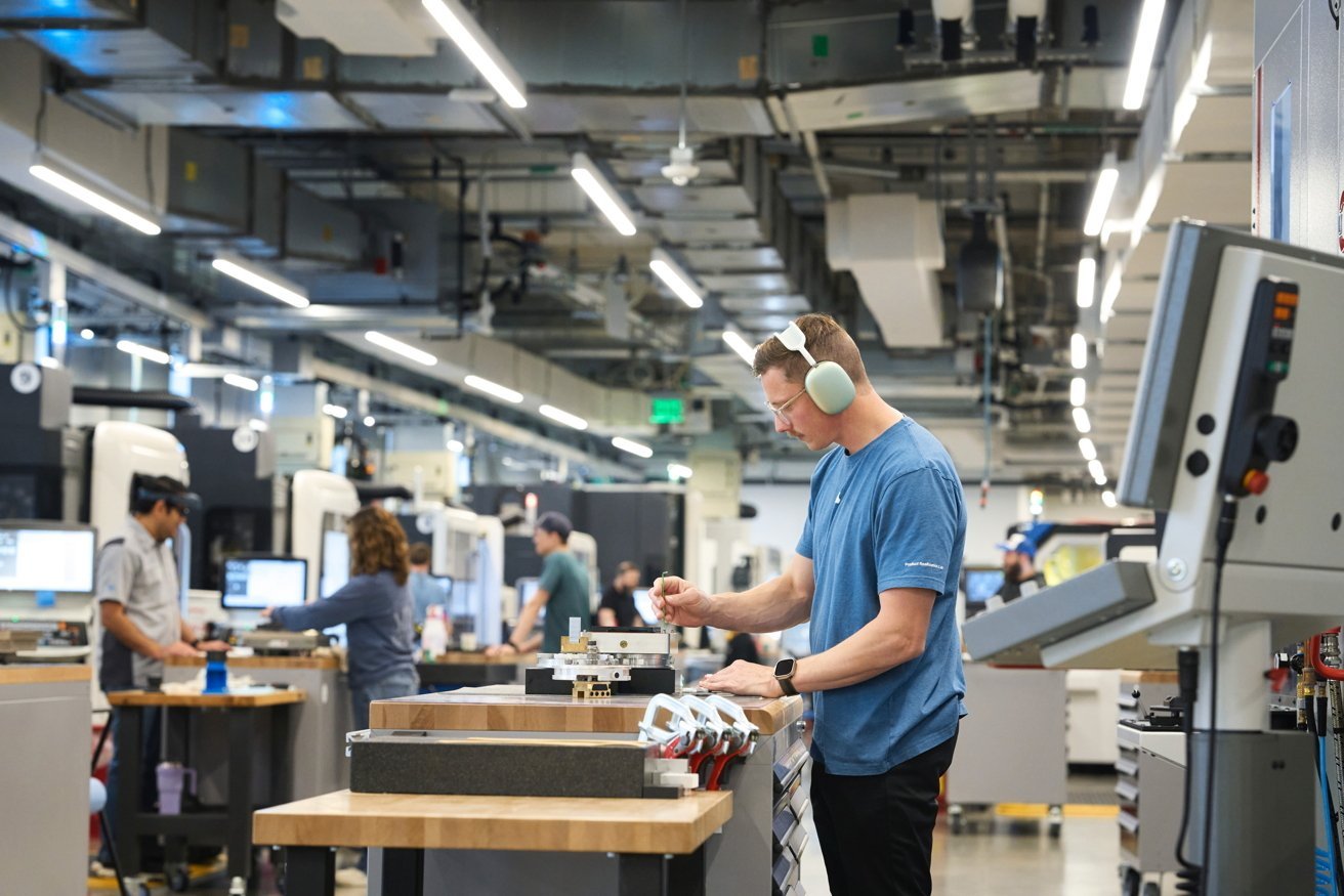 A busy factory floor with people working at tables, wearing casual clothes and protective earmuffs, surrounded by machinery and computers under bright overhead lighting.