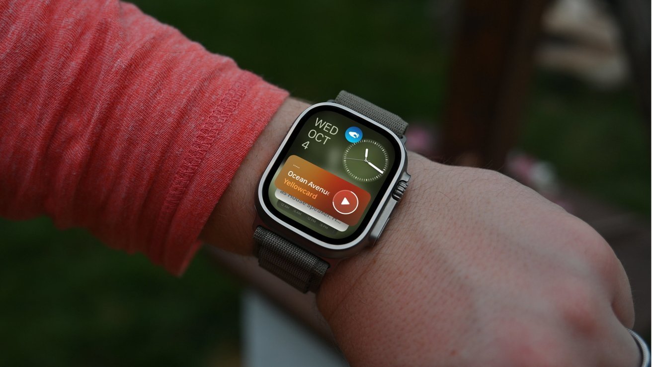 Person wearing a smartwatch displaying the time, date, and playing a song. The wearer is in a red long-sleeve shirt, and the watch has a fabric strap.