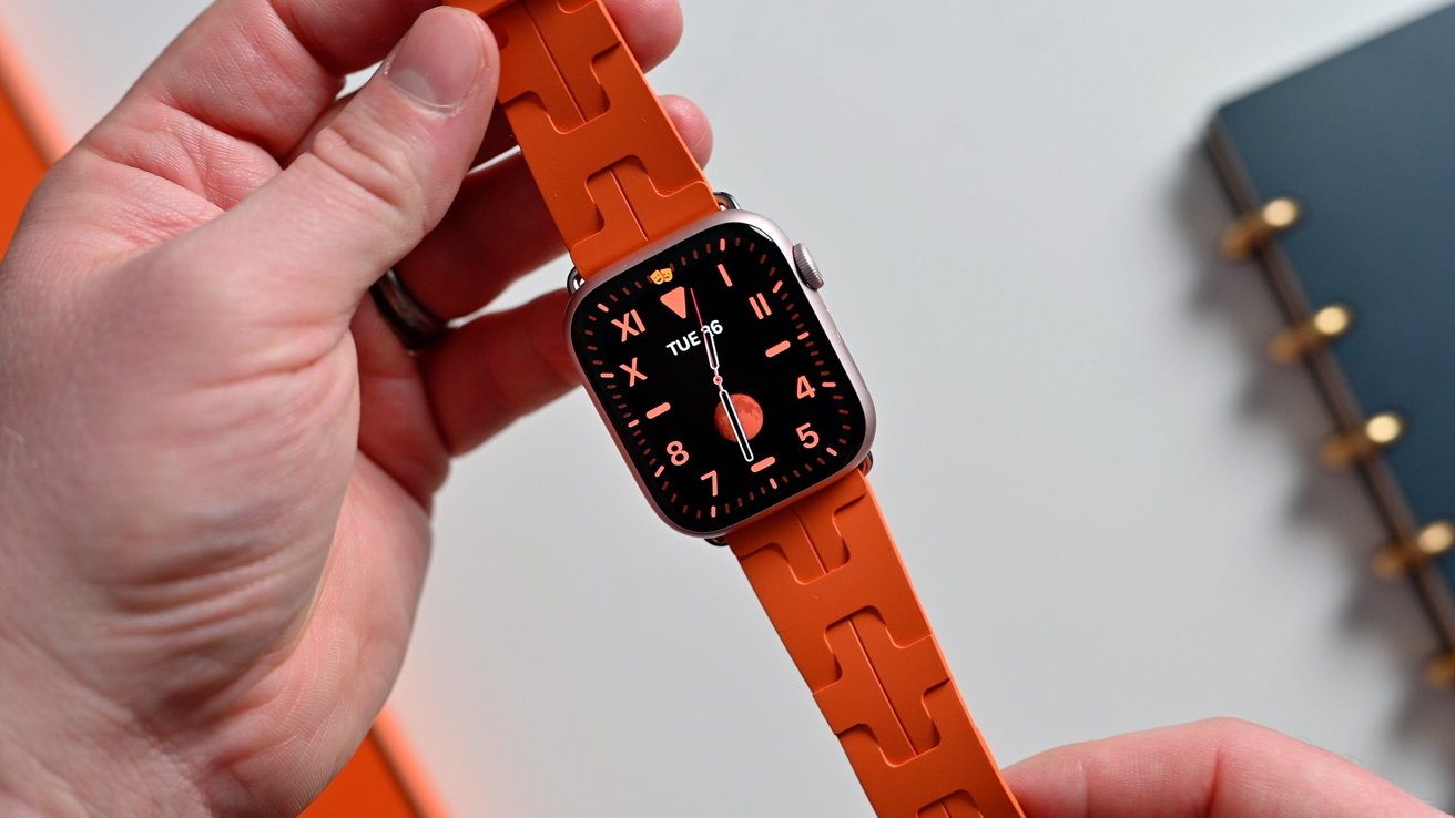A hand holding an Apple Watch with an orange band, displaying an analog watch face with Roman numerals and the date 'Tue 26'.