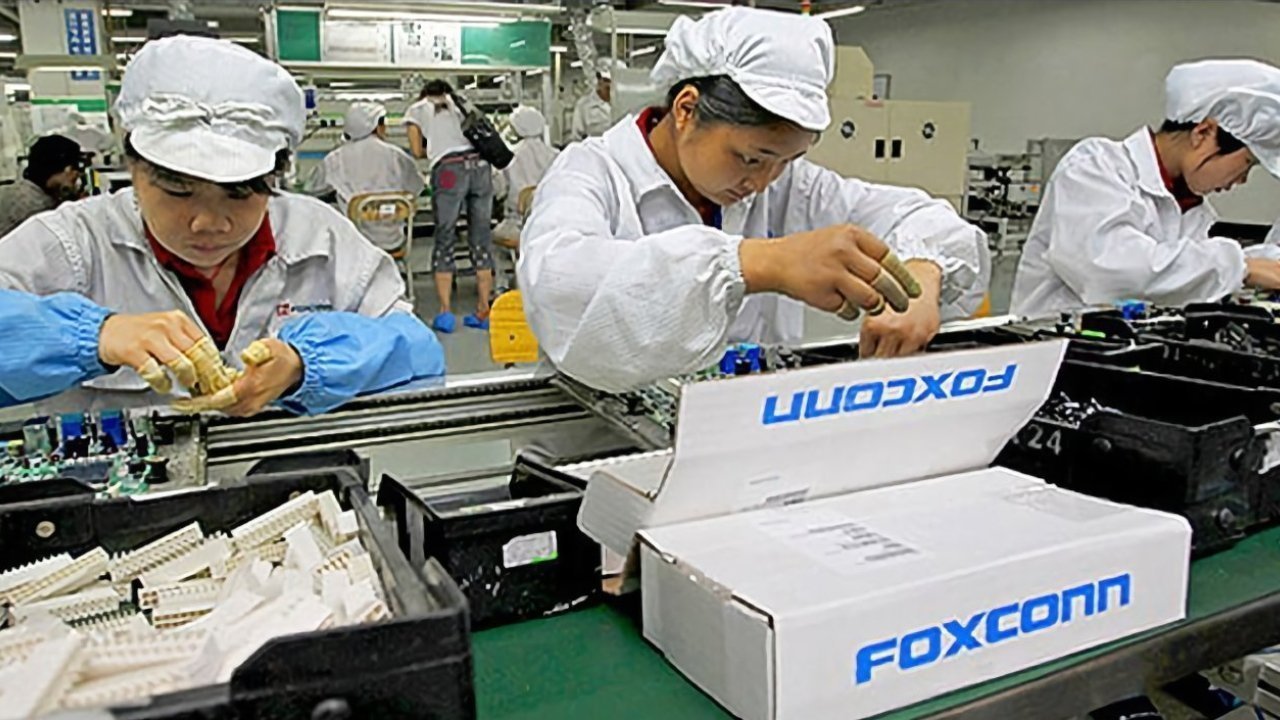 Workers in white uniforms assemble electronics at a factory workstation with boxes labeled Foxconn.
