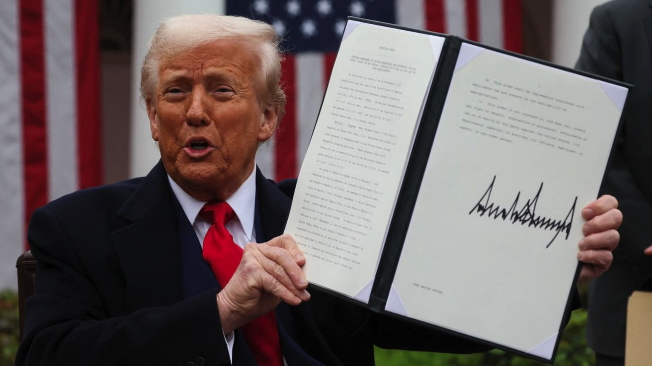 Man in a suit holds an open folder with a signed document, speaking with an American flag in the background.