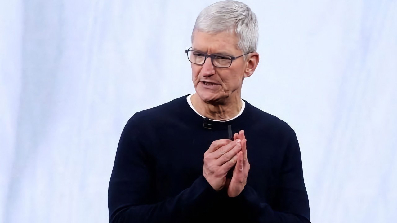 Man with gray hair and glasses, wearing a black shirt, clasping hands, speaking against a plain light background.