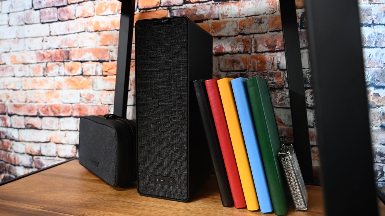 A speaker, colorful books, and a small black case are arranged on a wooden shelf against a red brick wall.