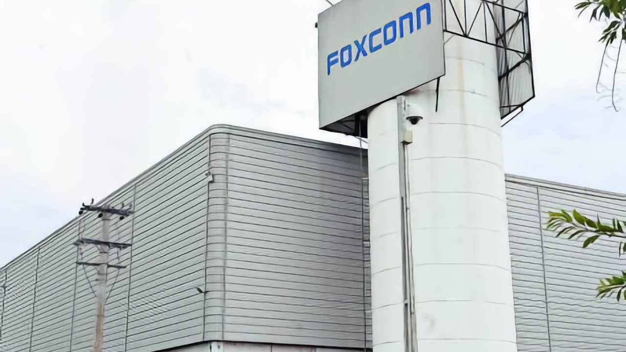 Gray industrial building with a large white column, displaying the 'Foxconn' logo in blue letters. Sparse greenery and a utility pole are visible. Overcast sky in the background.